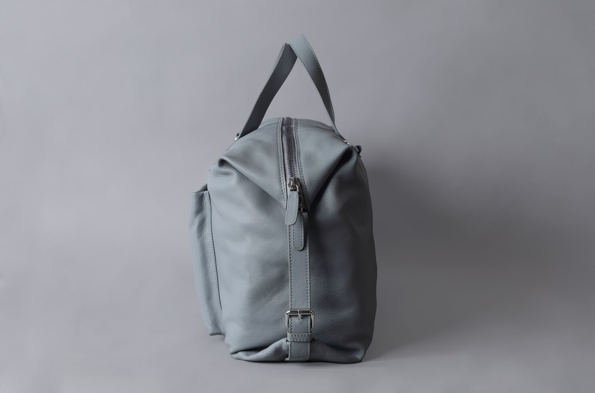 A light gray leather duffle bag with a zipper and handle, set against a plain gray background.