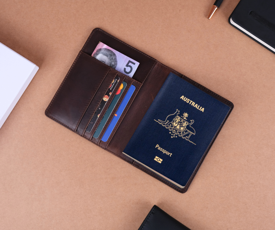 A brown leather passport holder open on a beige surface, showing an Australian passport, a five-dollar note, and several cards.