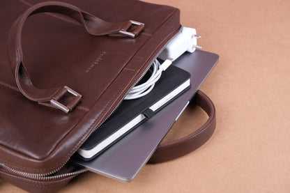 A brown leather bag is partially open, showing a laptop, a notebook, a charging cable, and a power adapter inside, against a brown background.