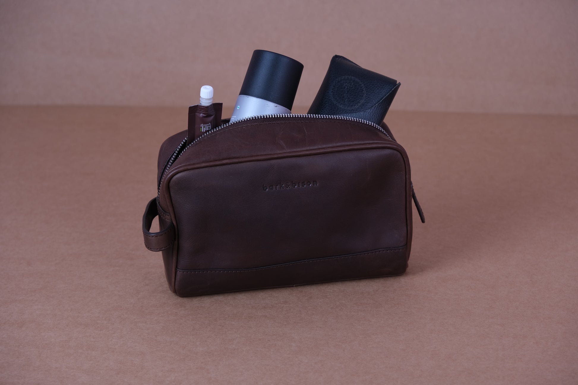 A brown leather toiletry bag is open, revealing a bottle, a tube, and a folded item inside.