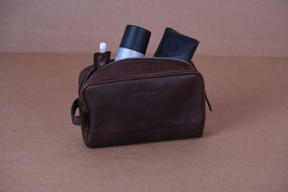 A brown leather toiletry bag is open, revealing a bottle, a tube, and a folded item inside.