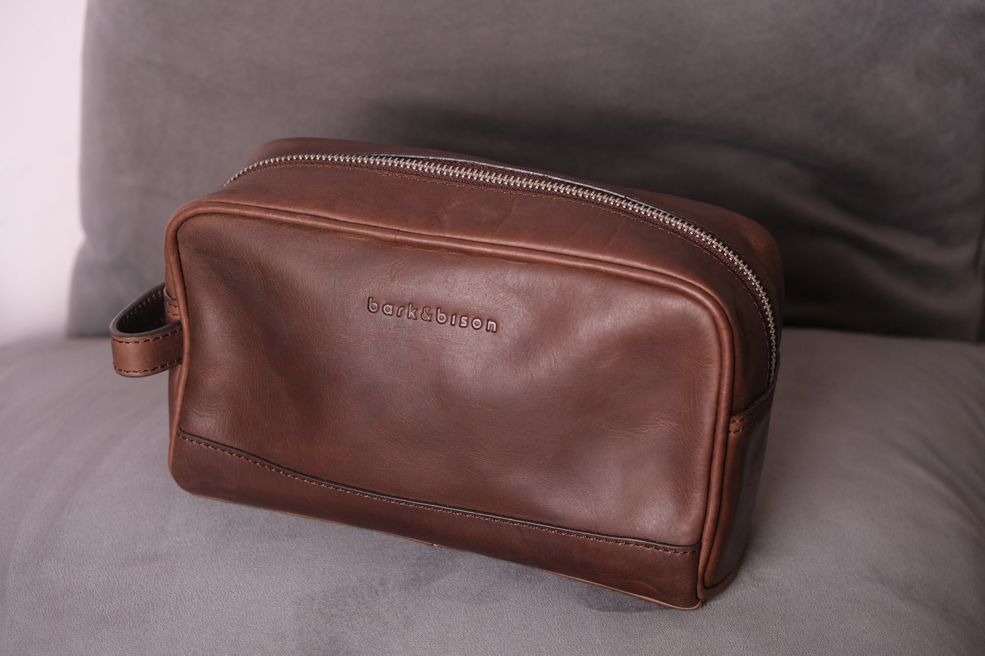 a brown leather toiletry bag with zipper and side handle kept on sofa embossed with bark&bison 