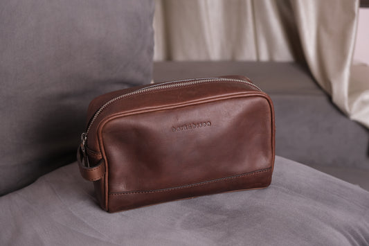 a brown leather toiletry bag with zipper and side handle kept on sofa embossed with bark&bison 