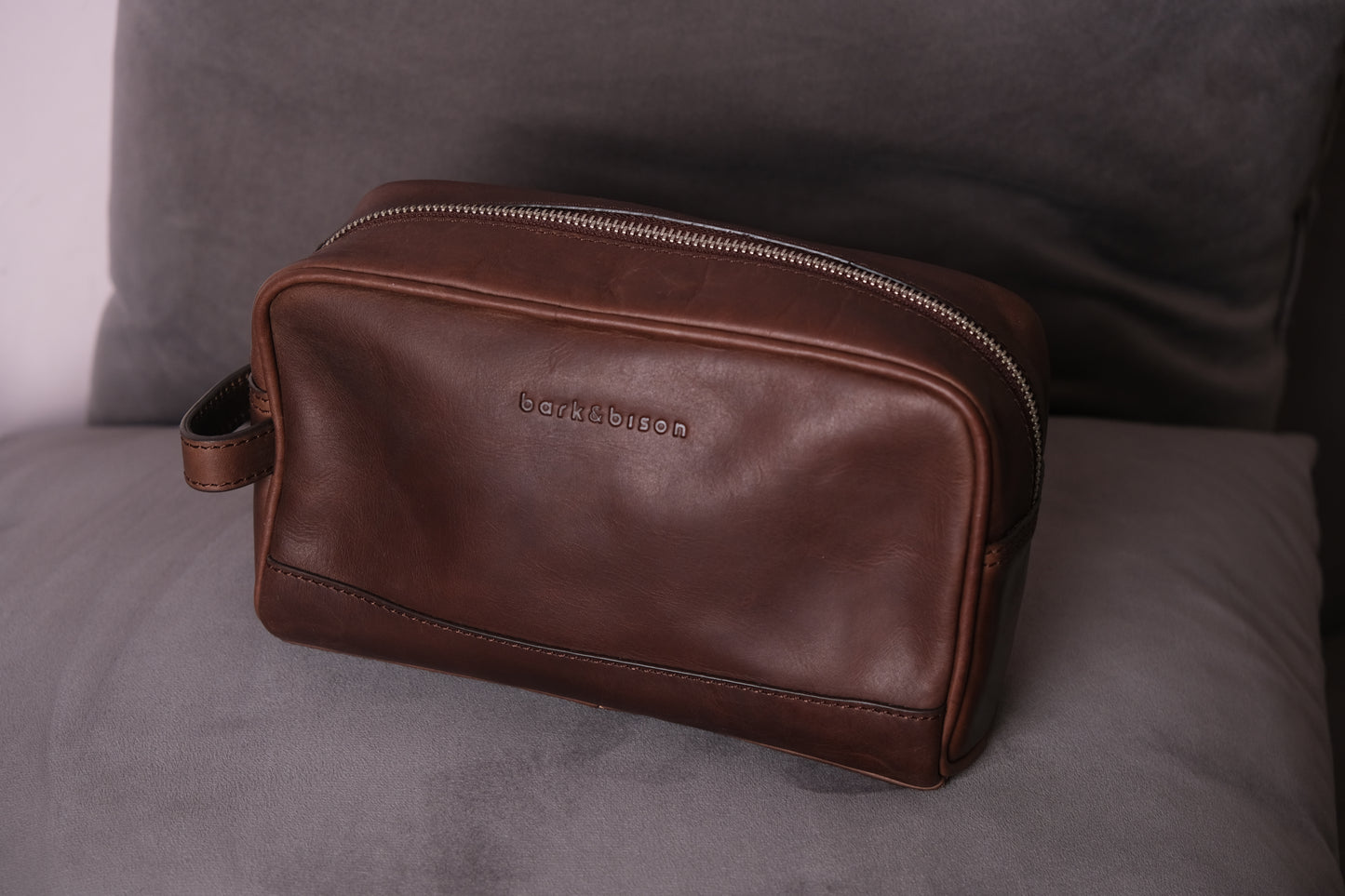 a brown leather toiletry bag with zipper and side handle kept on sofa embossed with bark&bison 