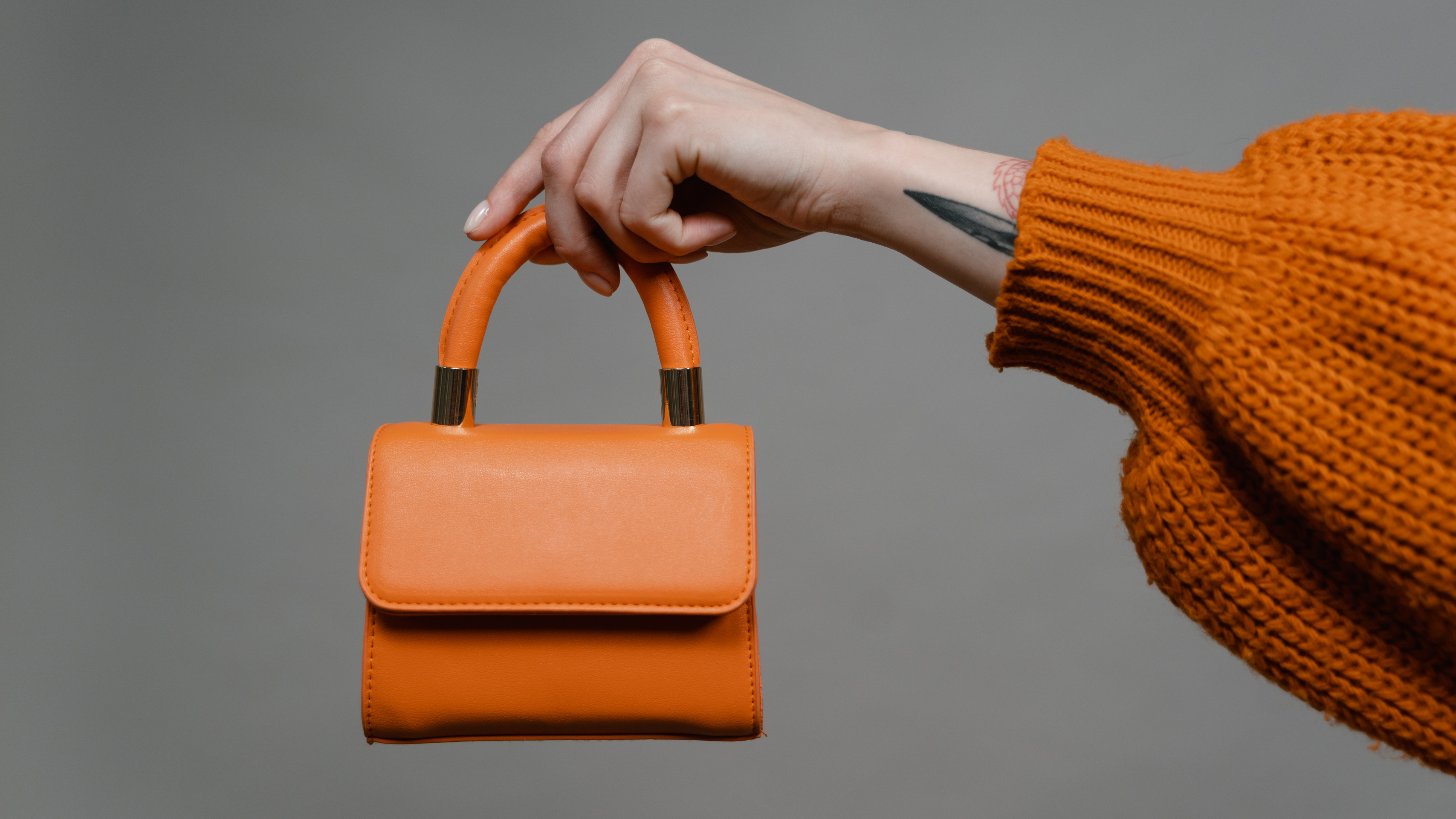 A hand gracefully holds an orange handbag with a gold accent against a gray background. The person complements the look with an orange sweater.