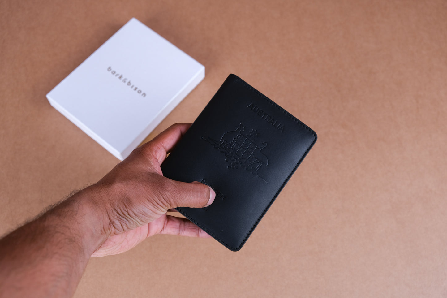 A hand holding a black Australian passport cover embossed with the national emblem, with a white box labeled "bark&bison" on a beige background.