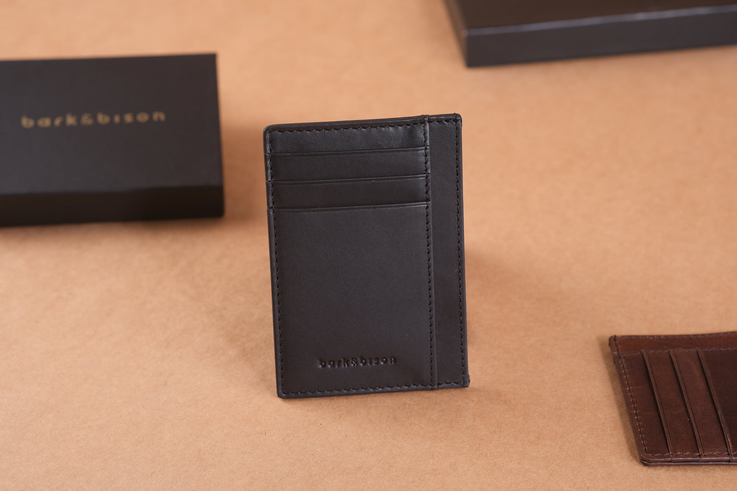 A black leather cardholder with several slots is displayed upright on a brown surface next to a black box with bark&bison branding.