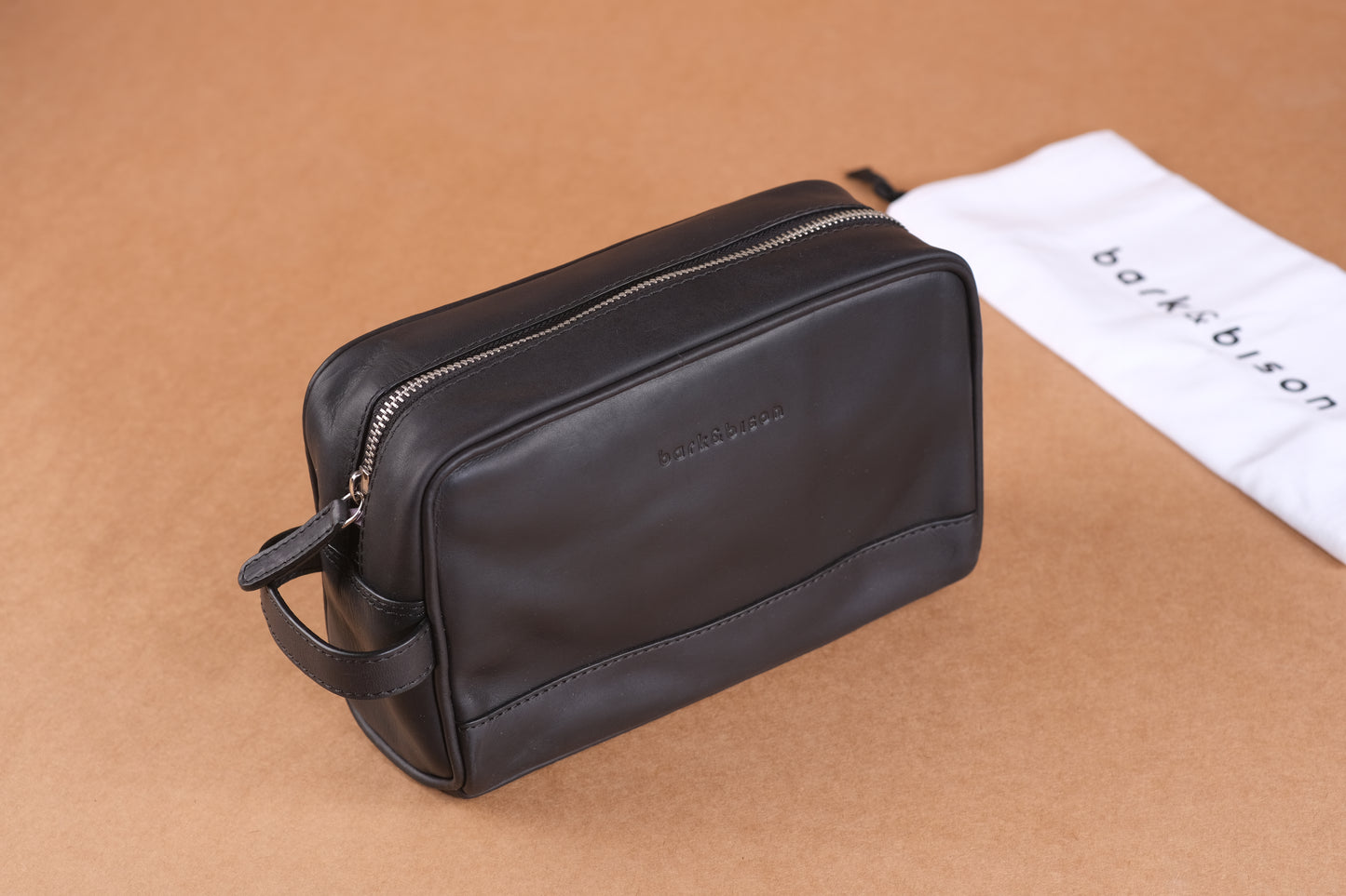 A black leather toiletry bag with a zipper, embossed with "bark&bison," displayed on a beige surface, with a white branded drawstring bag in the background.