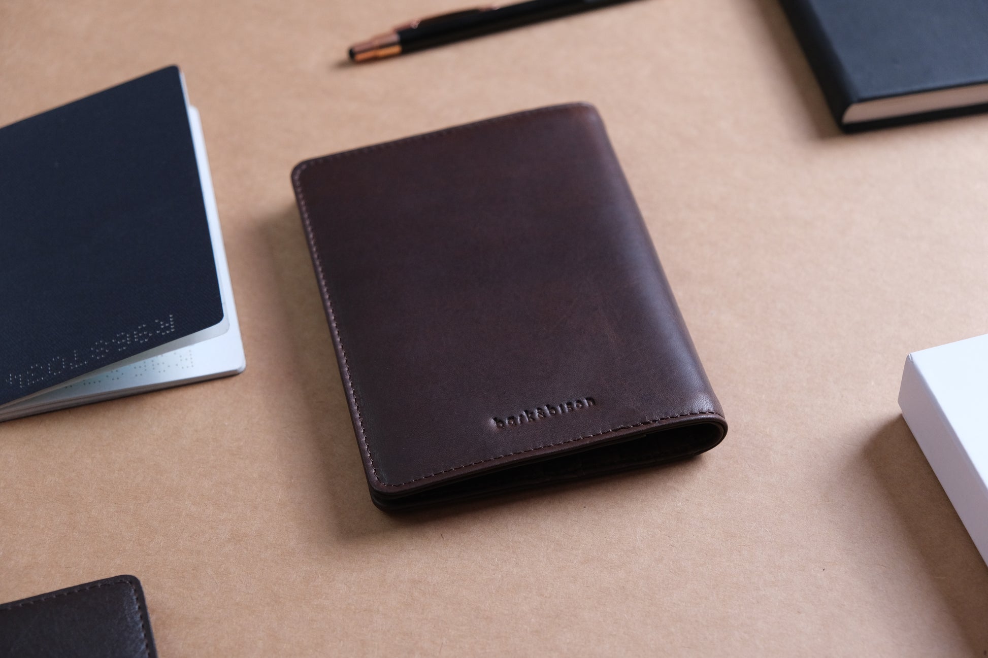 Brown leather passport holder with embossed Australian coat of arms and "Passport" text, partially holding a  passport. Positioned on a beige surface alongside a white "bark&bison" box, black wallet, and a black pen with rose gold accents.