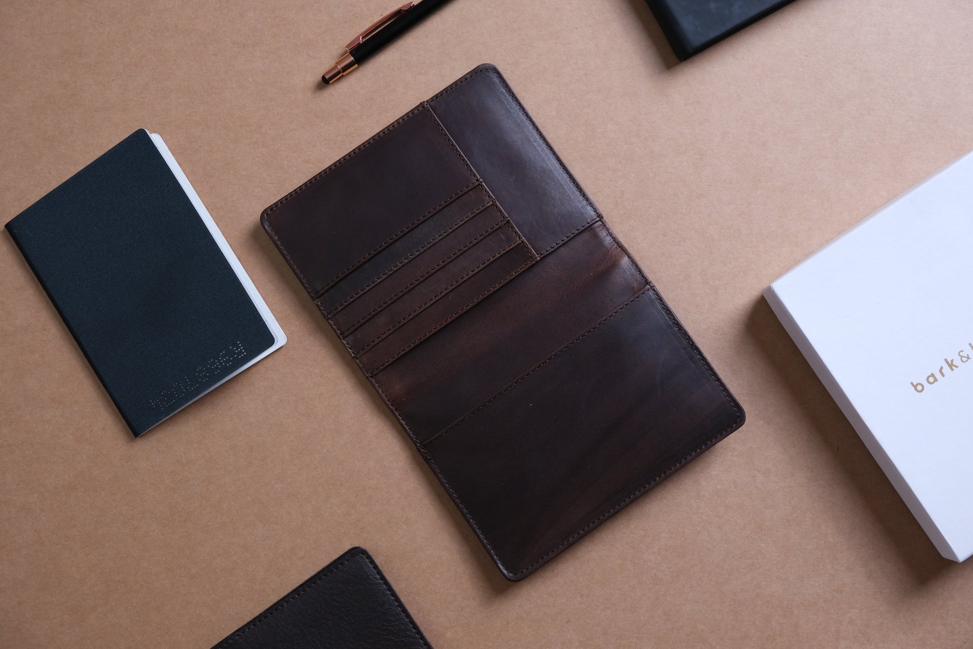 A brown leather passport holder with multiple slots open next to a passport and a pen with a white box embossed with bark&bison branding.