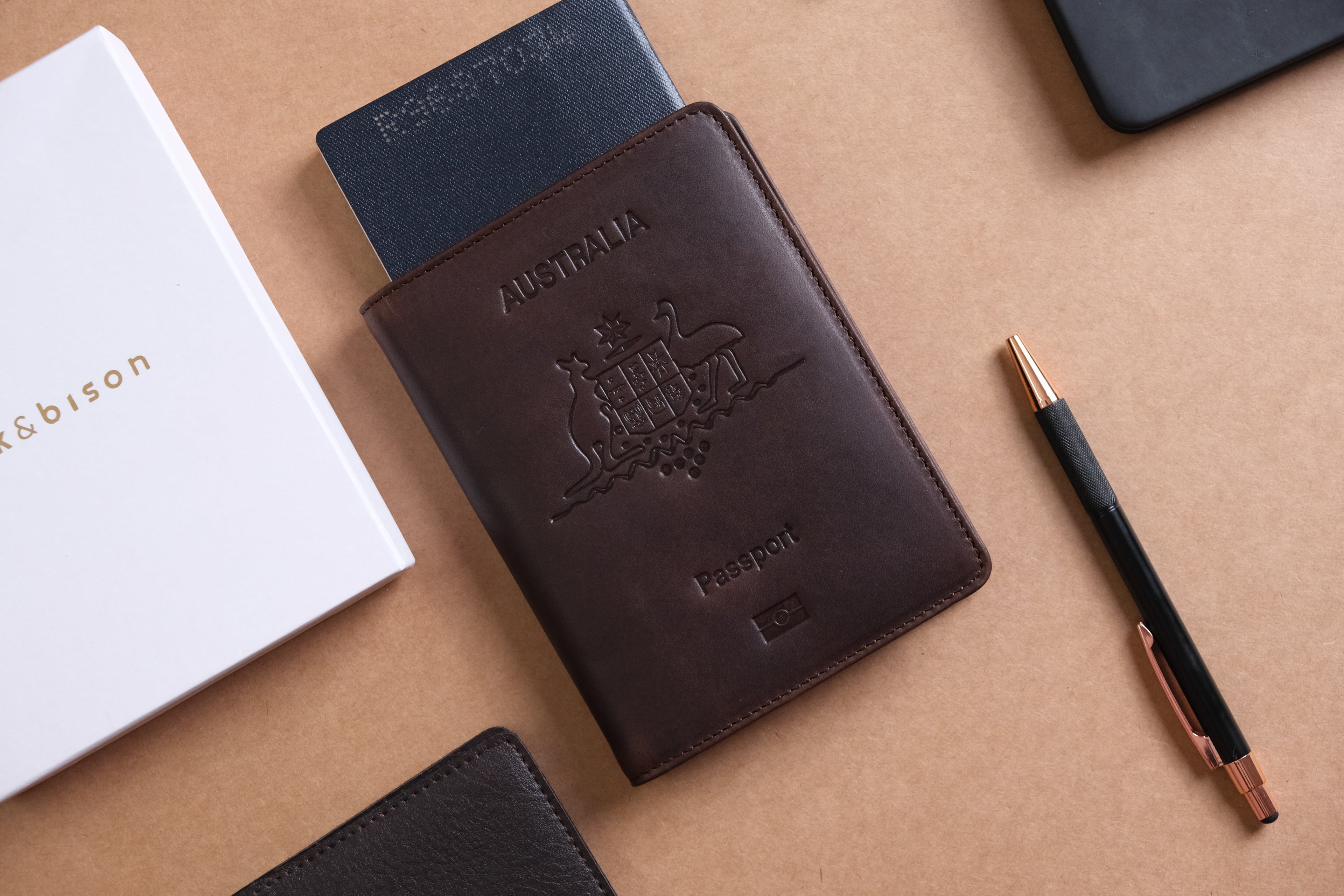 Brown leather passport holder with embossed Australian coat of arms and "Passport" text, partially holding a  passport. Positioned on a beige surface alongside a white "bark&bison" box, black wallet, and a black pen with rose gold accents.