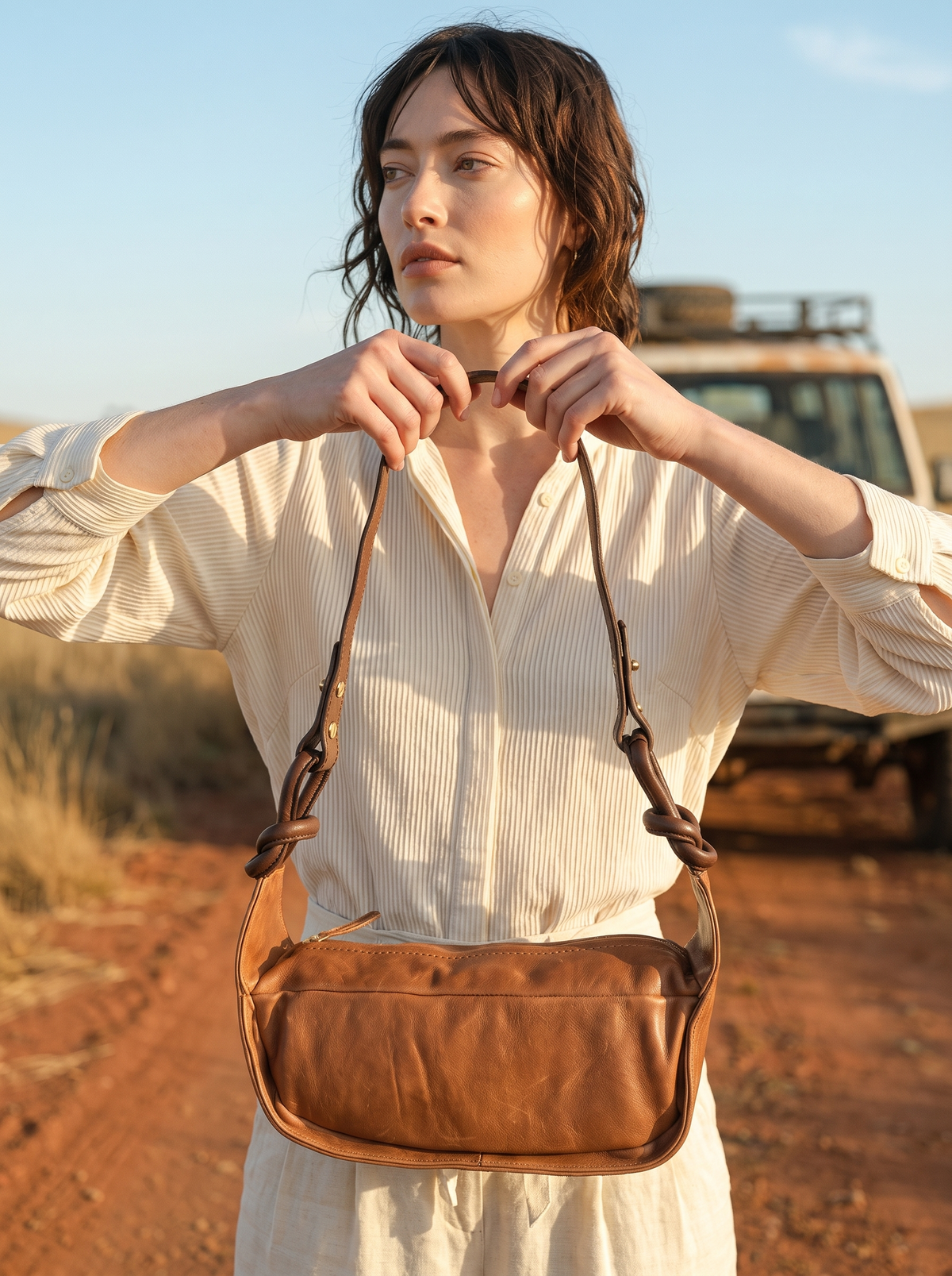 Model holding the Pilbara Knot chocolate leather hobo bag by its braided handle, with a 4WD in the background