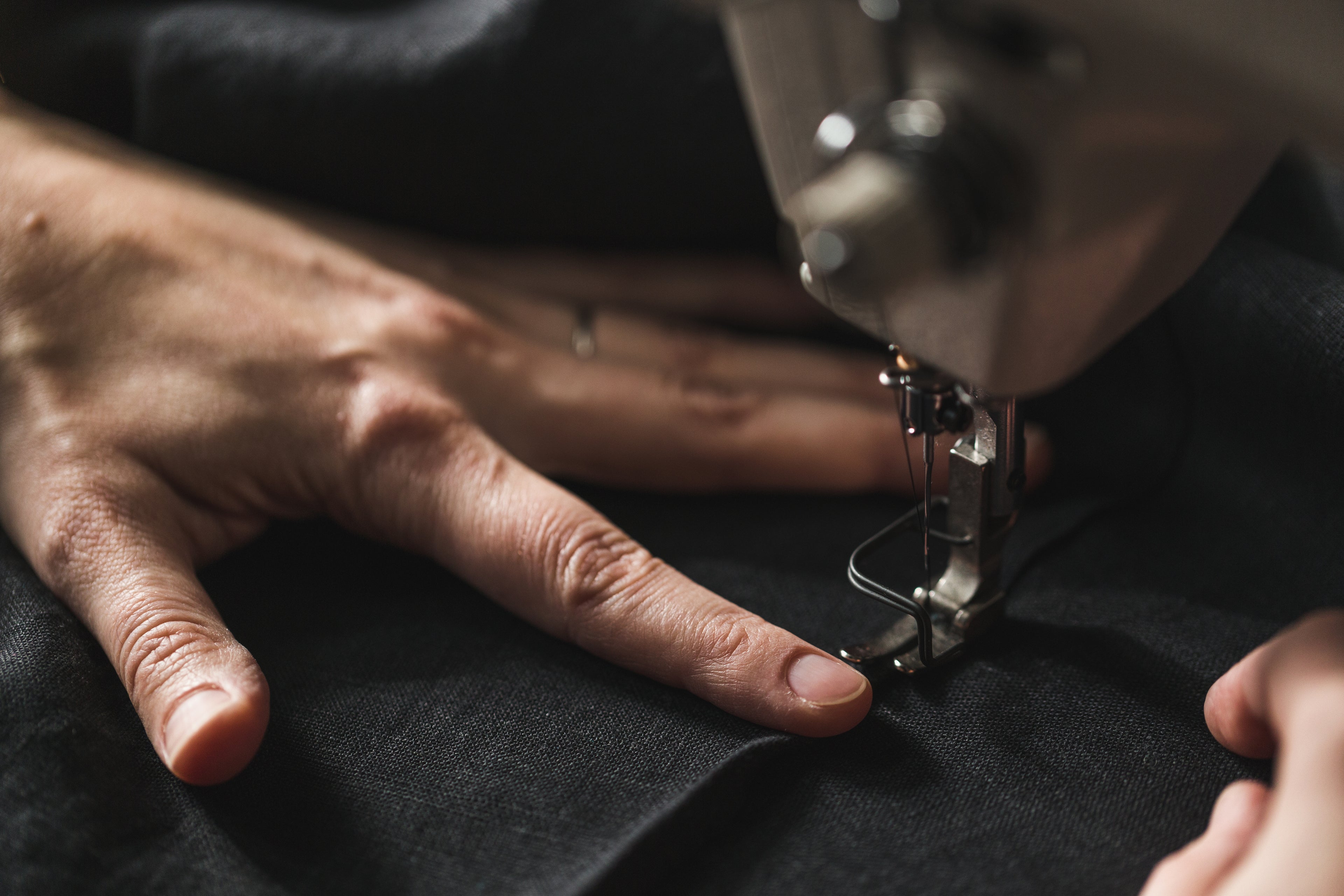 Close-up of a person’s hand guiding a piece of dark fabric through a sewing machine. The sewing machine needle is positioned near the fabric, indicating active stitching or tailoring. The scene highlights precision craftsmanship.