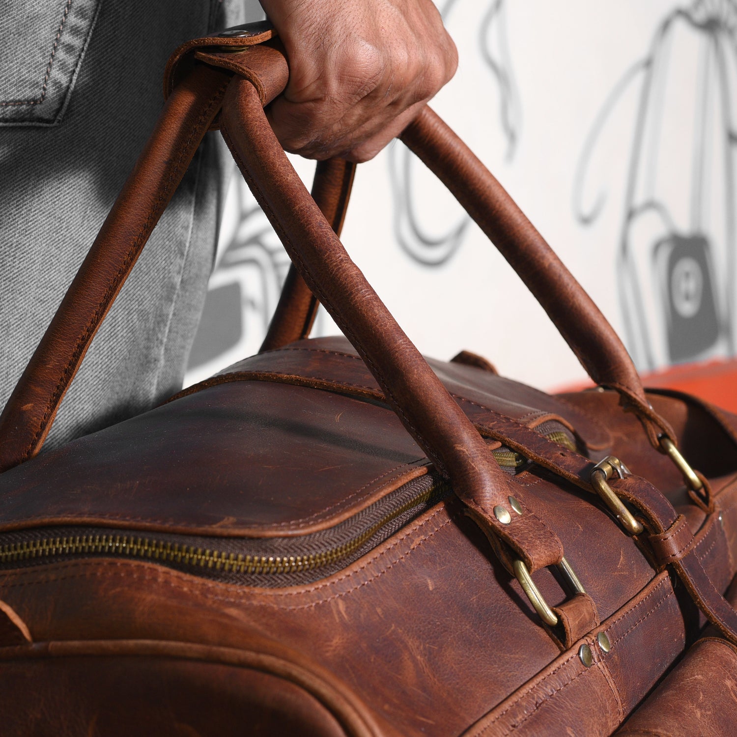 Close-up of a person holding a brown leather bag by its handle, with a gray and white background.